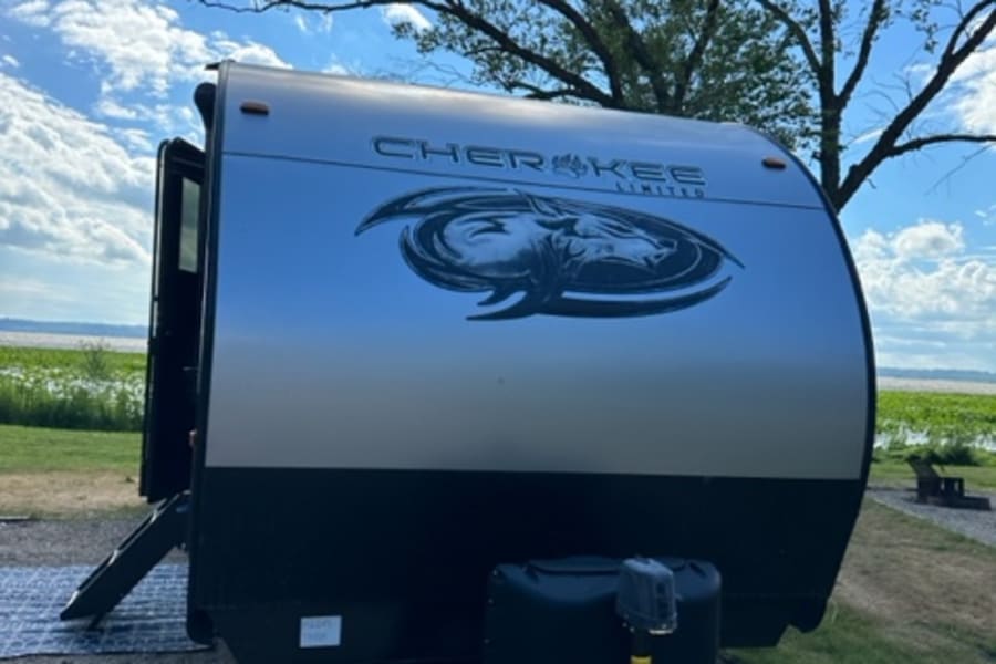 RV rental: A white and black Cherokee Limited travel trailer parked on gravel, with tree branches and a blue sky overhead.