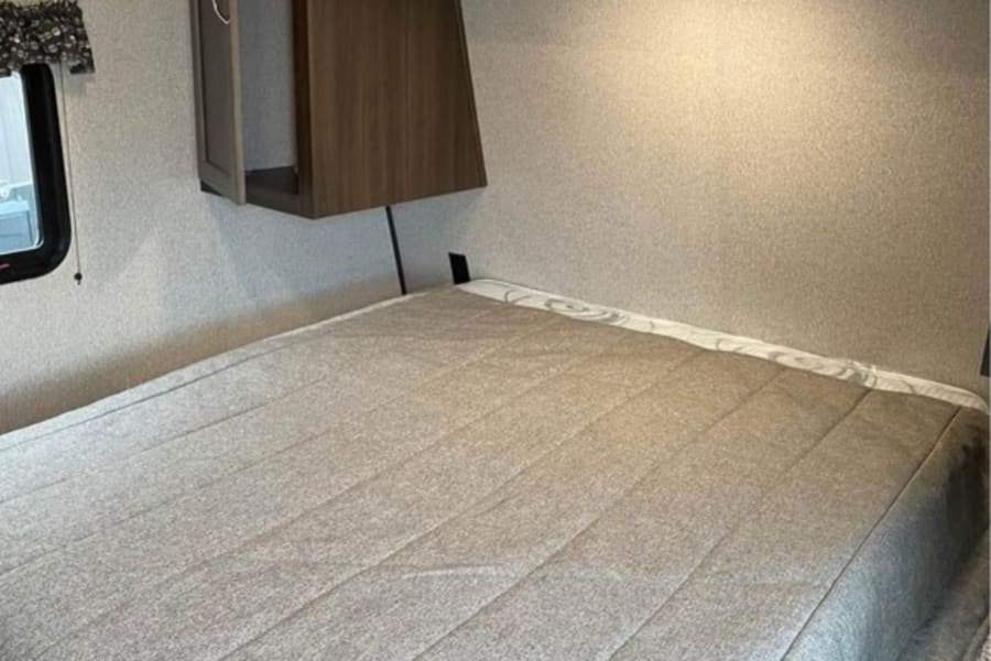 Cozy RV rental bedroom features a comfortable bed with grey bedding, overhead storage, and soft lighting for your travels.