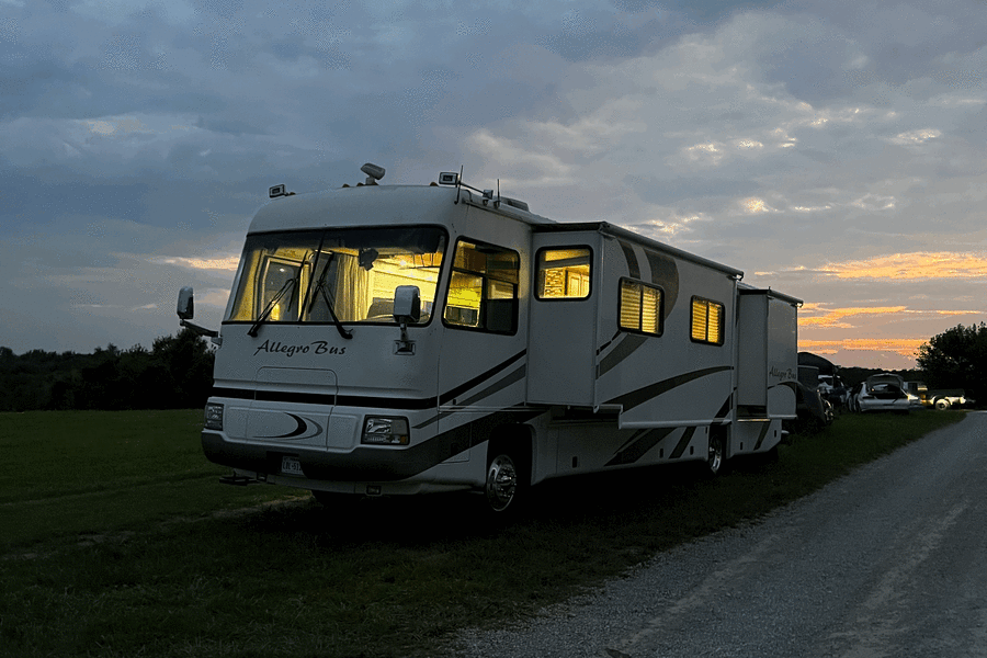 Cozy Allegro Bus RV rental bathed in interior light at dusk, parked on grass by a gravel road with a sunset sky.