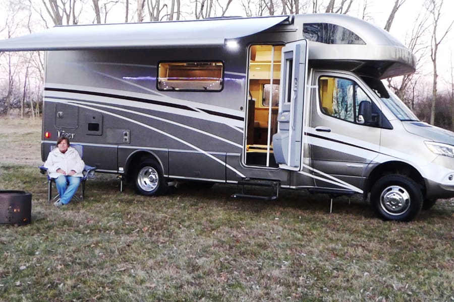 This RV rental features a sleek motorhome with an open door & extended awning. A person relaxes by a fire pit in a grassy outdoor setting.