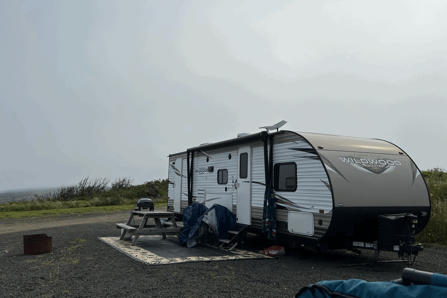 Wildwood rv rental travel trailer set up on a gravel camping site with picnic table, grill, and fire pit under misty skies.