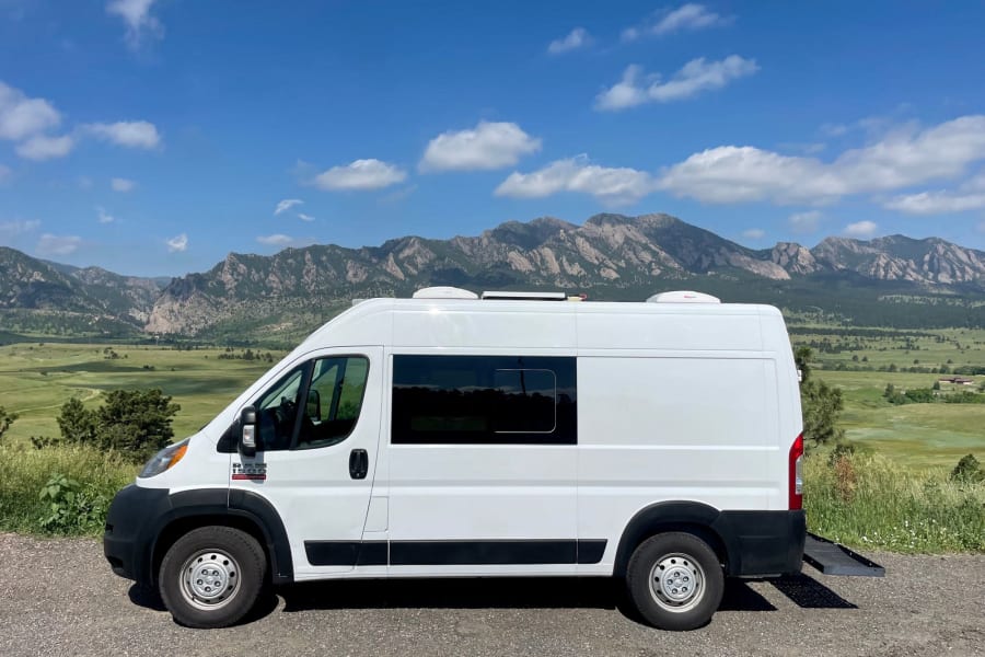 White camper van, likely an RV rental, parked on gravel with scenic mountains and blue sky in background. A ramp extends from the rear.
