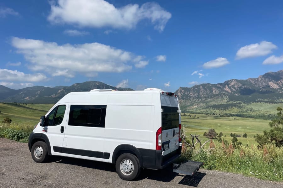 Scenic RV rental: A white camper van parked on a dirt road with green valleys & distant mountains under a blue sky.