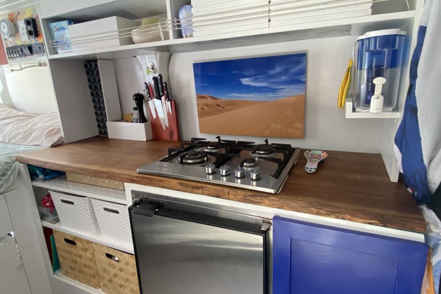 Inside an RV rental, a compact kitchen with wood-look counter, built-in stovetop, fridge, and storage. Perfect for road trips.
