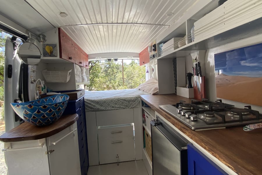 Inside this bright RV rental camper van: A full view of the kitchenette with stove, wooden counter, bed, and decorative blue sink.
