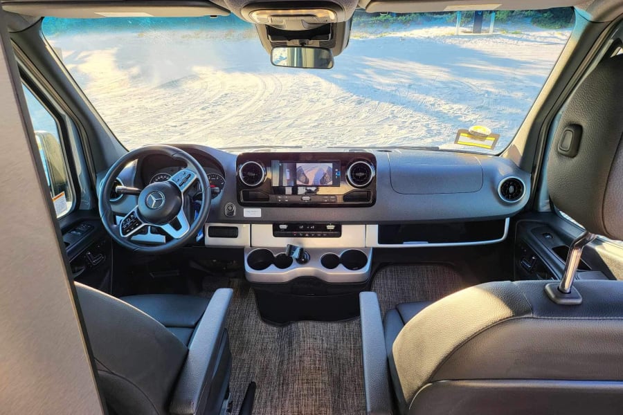 RV rental interior view: spacious driver's cockpit with dashboard, steering wheel, and front seats, parked on sandy terrain.