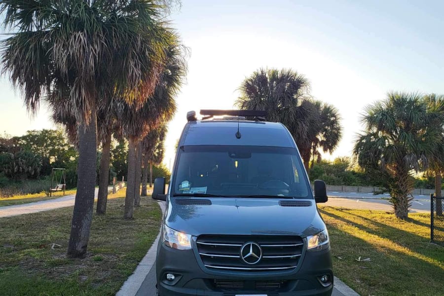 A sleek dark grey Mercedes Sprinter RV rental parked on a sunny road lined with palm trees. Perfect for adventure!