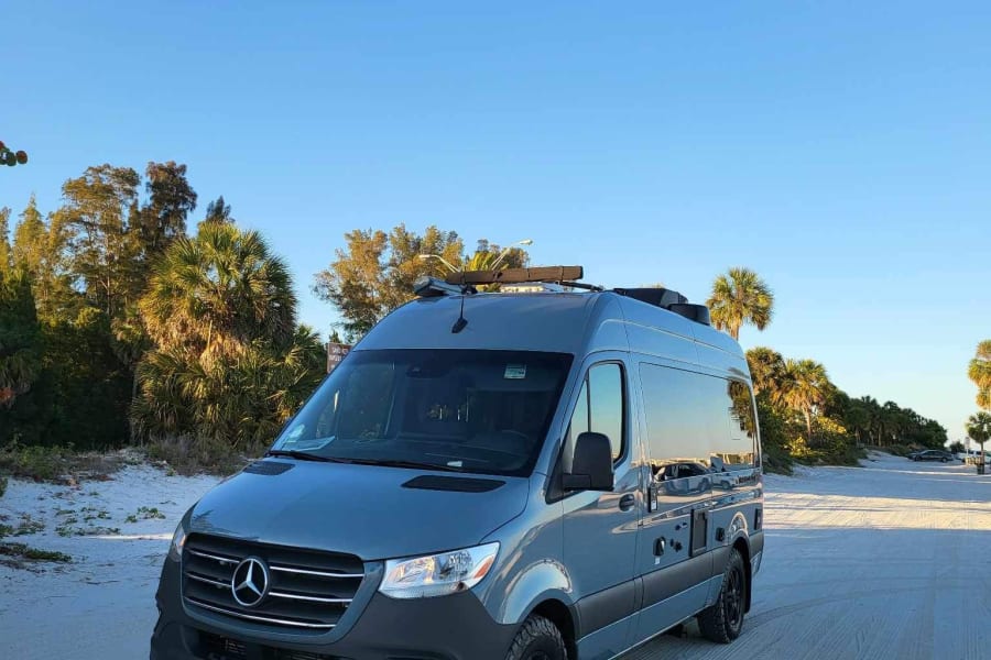Gray Mercedes Sprinter RV rental on a sandy beach road among palm trees under a clear blue sky.
