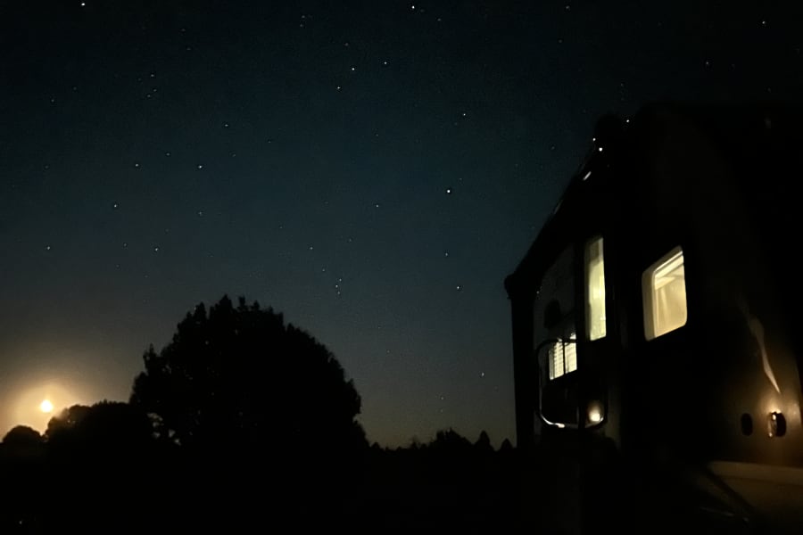 Looking up at a starry night sky from an RV rental, with interior lights on and silhouetted trees.