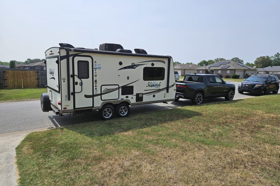 Green Rivian electric truck tows a white Shamrock travel trailer (RV), parked on a residential street, ideal for an rv rental.