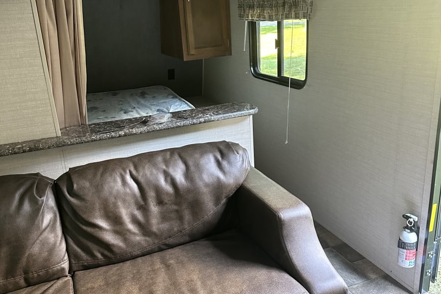 Interior of an RV rental featuring a brown leather sofa, bed area, overhead cabinets, and a mounted fire extinguisher.