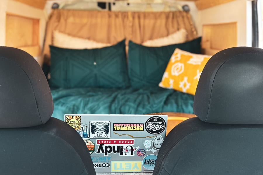 Cozy RV rental interior. View from front seats to bed with teal/yellow pillows. Wooden ceiling & laptop with stickers.