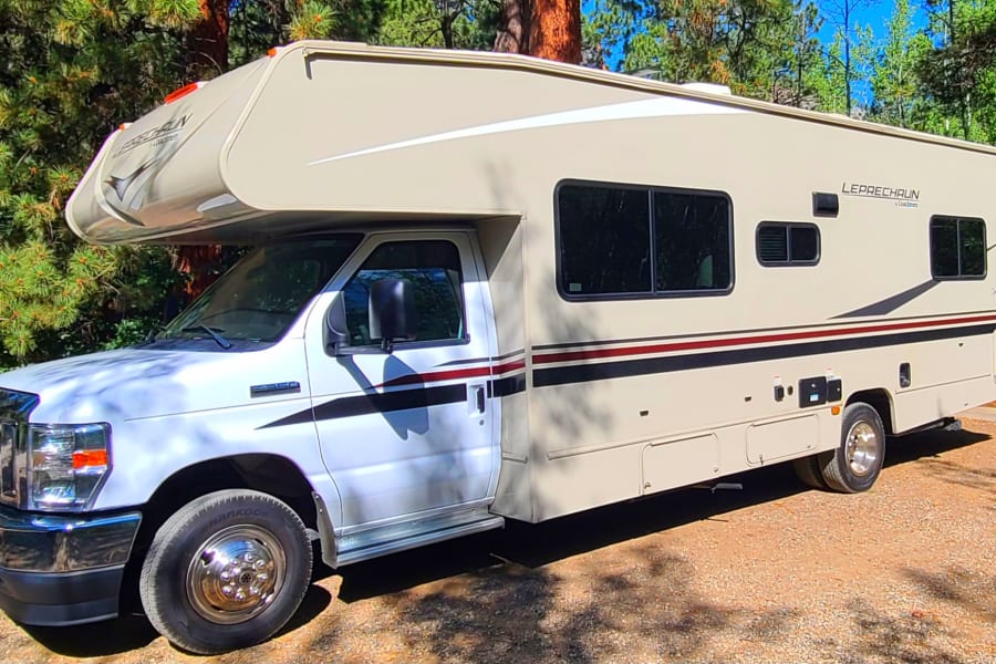 Spacious tan Class C **rv rental** parked on gravel, surrounded by green trees under a sunny sky.