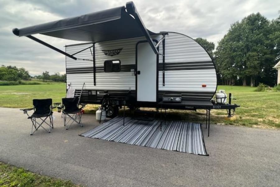 This RV rental is set up for outdoor comfort, featuring an extended awning, chairs, and a striped rug on an asphalt pad.