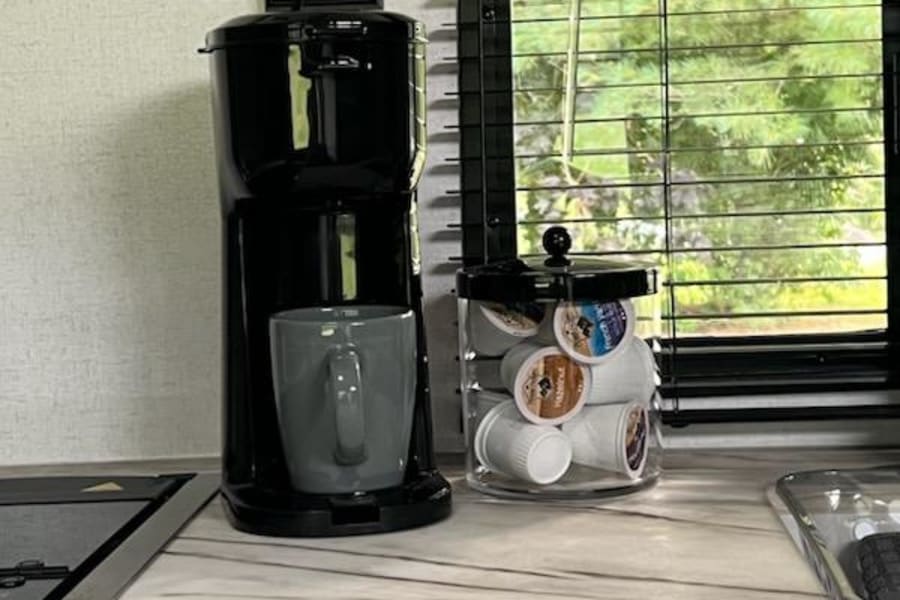 An RV rental features a convenient coffee station with a black maker, grey mug, and K-cup storage jar on the counter.