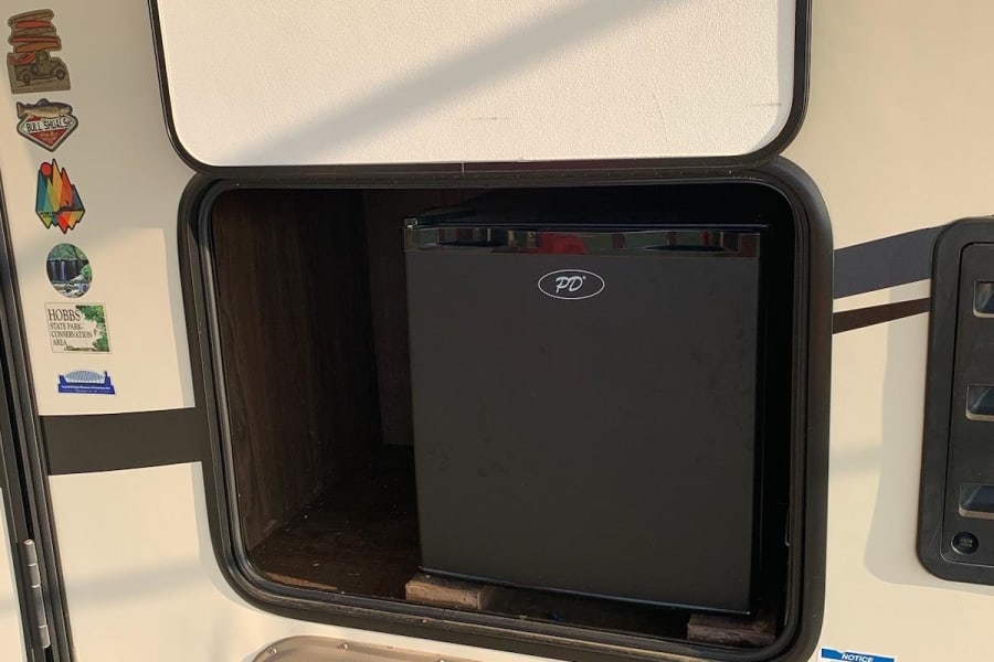 An RV rental's exterior storage hatch is open, showing a black compact refrigerator inside.