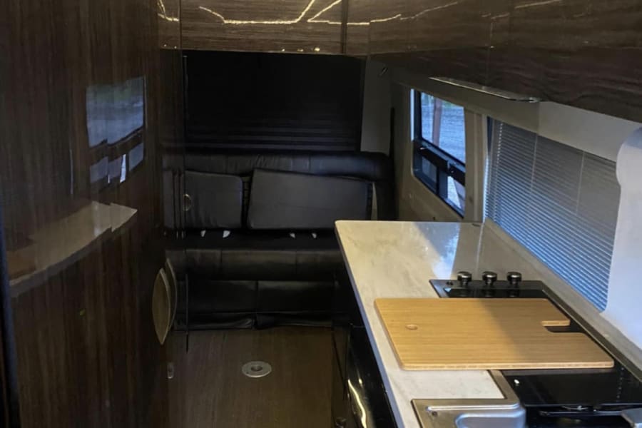 Modern RV rental interior: galley kitchen, comfortable rear lounge/bed, wood floors, and bright overhead LED lighting.