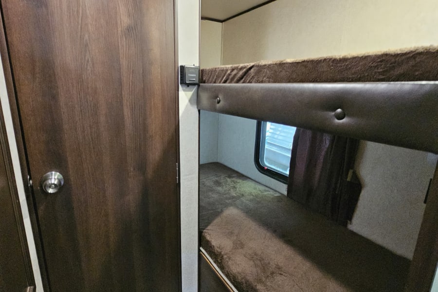 Inside an RV rental: a bunk bed with a window, dark wood door with handle, and a Dometic control panel.