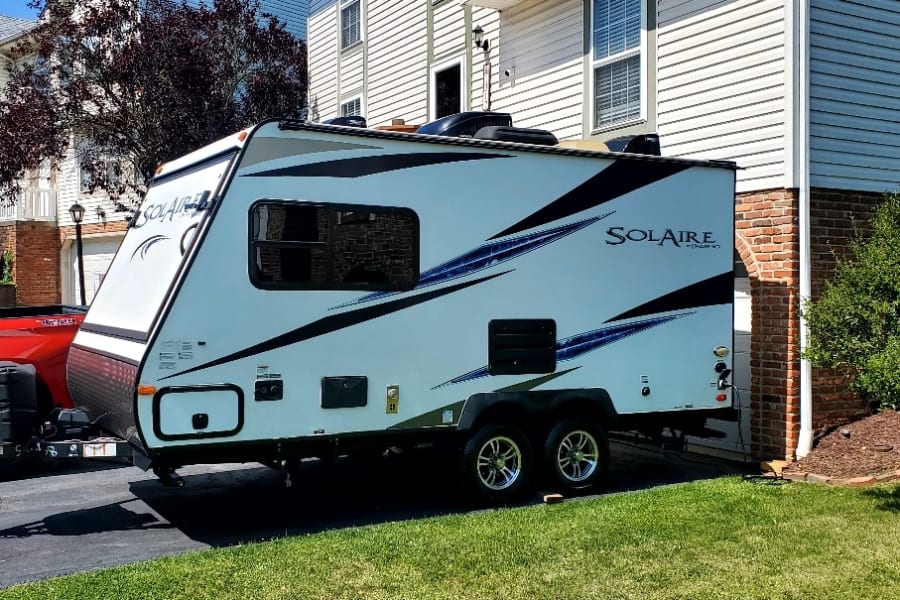 White Solaire travel trailer, ready for an RV rental, parked on a residential driveway connected to a red truck.