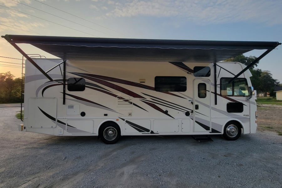 This white RV rental, parked on gravel with its black awning extended, offers comfort for your travels. Explore the open road!