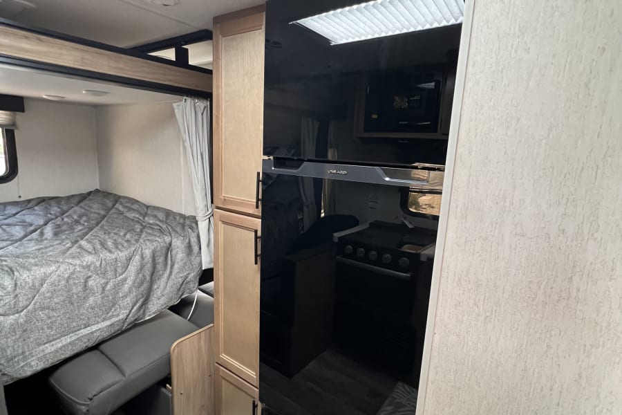 Cozy RV rental interior with a comfortable bed, pull-out bench, modern black refrigerator, and wood cabinets.