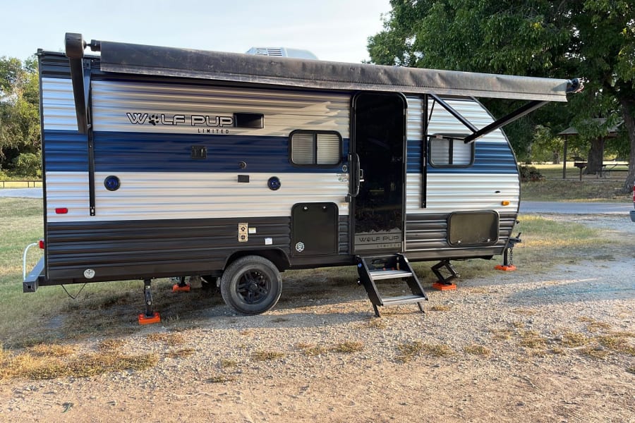 A blue and white Wolf Pup travel trailer RV rental is parked at a gravel campsite. Awning extended, steps down.