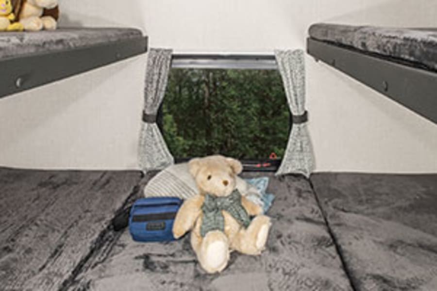 Inside an RV rental, two sets of comfy bunk beds with grey bedding & playful stuffed animals. Window views trees outside.