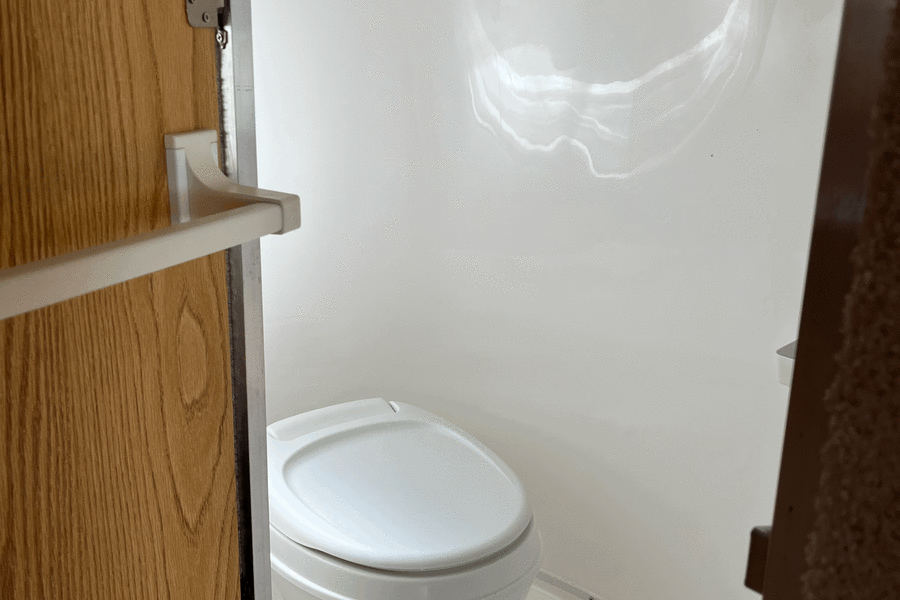 The clean, compact white toilet and integrated shower pan of an **rv rental** bathroom. A light wooden door is open on the left.