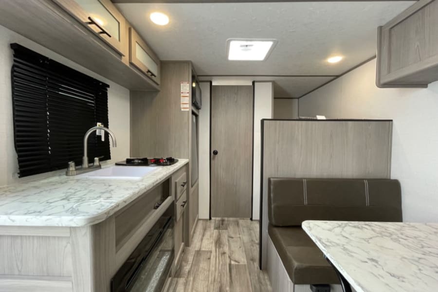 Modern RV rental interior showing a clean kitchen with marble-look counters, sink, and a cozy dinette with brown seating.