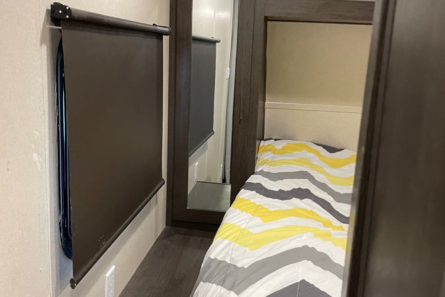 This RV rental bedroom offers a cozy bed with a yellow and gray striped comforter, dark window blind, and built-in storage.