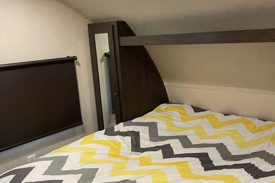 An RV rental bedroom boasts a bed with a yellow & gray chevron comforter, practical window blind, and built-in storage.