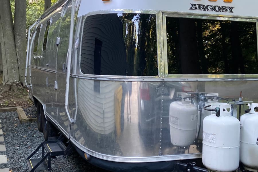 Silver & white Airstream Argosy RV rental with propane tanks, nestled on gravel in a peaceful, wooded setting.
