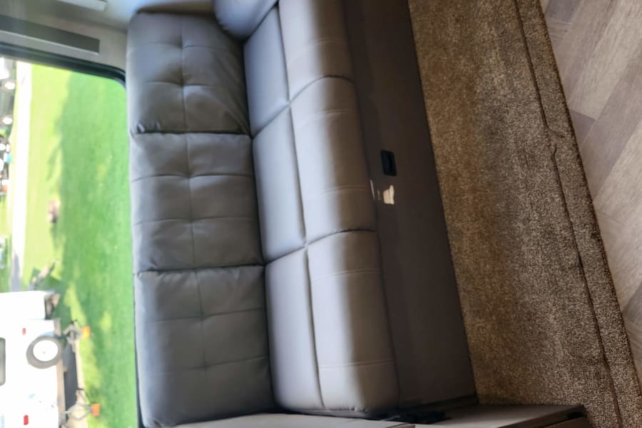 Cozy RV rental interior with tufted gray seating, plush brown carpet, and light wood-look flooring, revealing green outdoors.