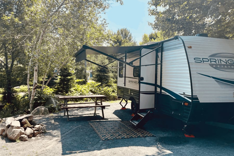 A white Springdale RV rental is parked at a sunny gravel campsite with a picnic table, fire pit, and extended awning.