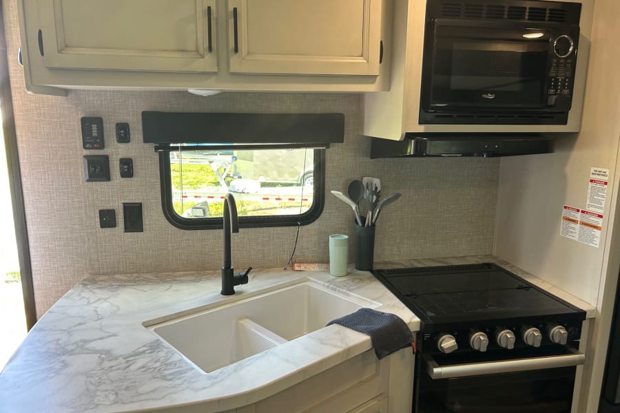 Inside an rv rental: Well-equipped kitchen with marble-look counter, double sink, black stove, microwave, and overhead cabinets.