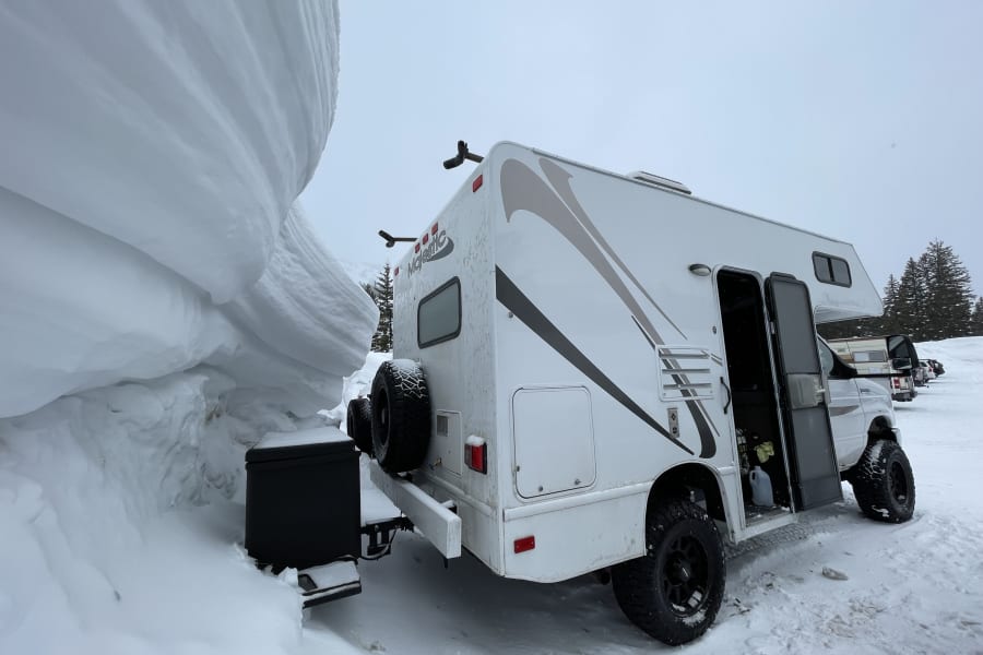 This rugged RV rental is dwarfed by a towering snowdrift, showcasing extreme winter camping in deep snow.