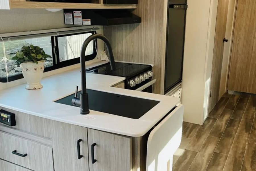 View of a clean, modern RV rental kitchen featuring a sink, stove, light wood cabinets, and dark flooring.