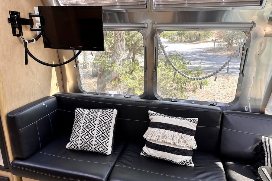 Inside an RV rental, a black leather sofa with patterned pillows faces a mounted TV. Large windows offer a view of trees and a road.