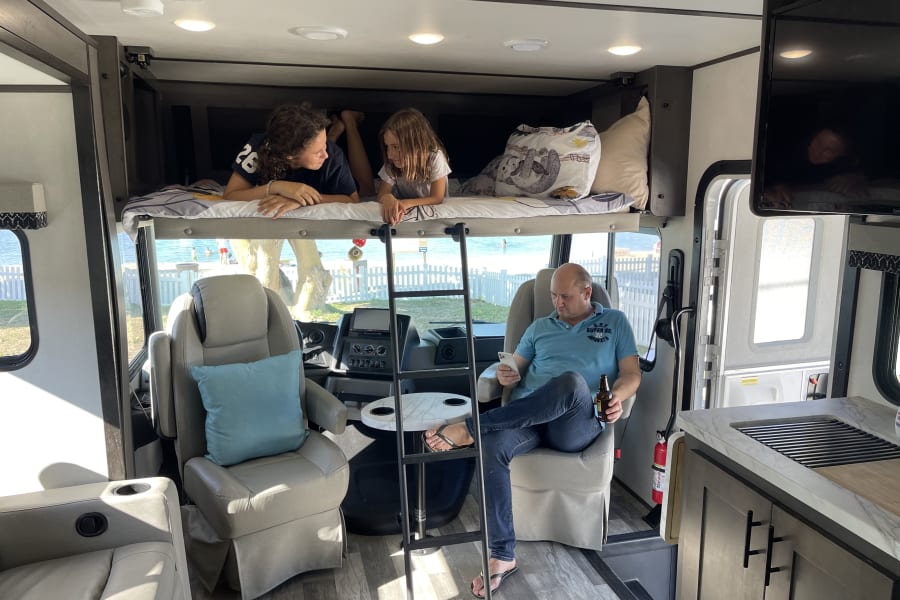 Relaxing in an RV rental: Kids chatting on the overhead bunk while dad unwinds with a drink, beach visible.