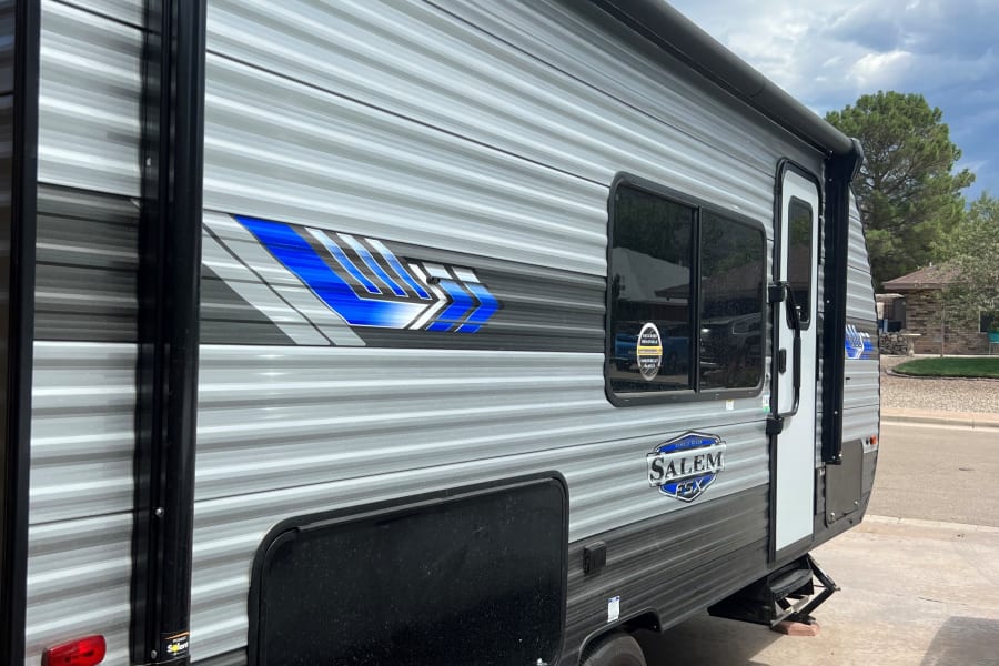 Gray RV rental travel trailer with blue accents, parked on a concrete driveway under a cloudy sky.
