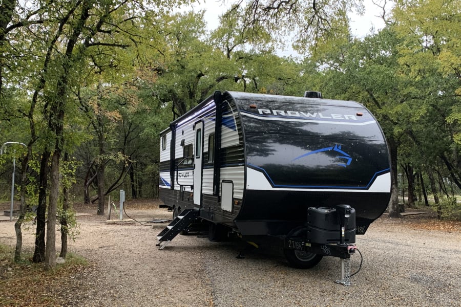 A black Prowler travel trailer RV rental is set up in a beautiful, wooded nature campsite.