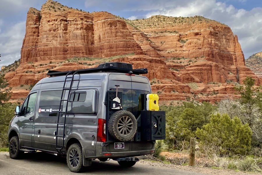 Grey RV rental van parked on road. Stunning red rock formations rise behind it under a cloudy sky. Explore adventure travel!