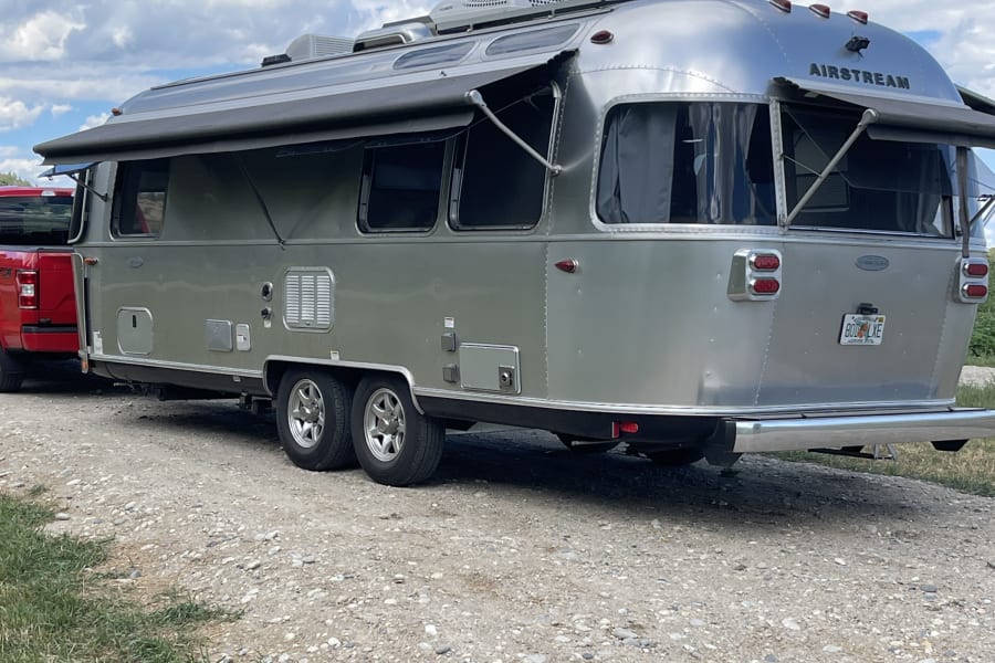 Silver Airstream RV rental hitched to a red truck, parked outdoors with its awning extended under a blue sky.