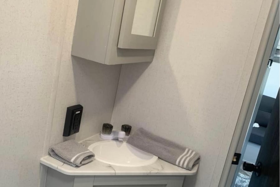 Compact corner sink area in an RV rental bathroom, featuring a grey vanity, mirror cabinet, wall outlet, and grey towels.
