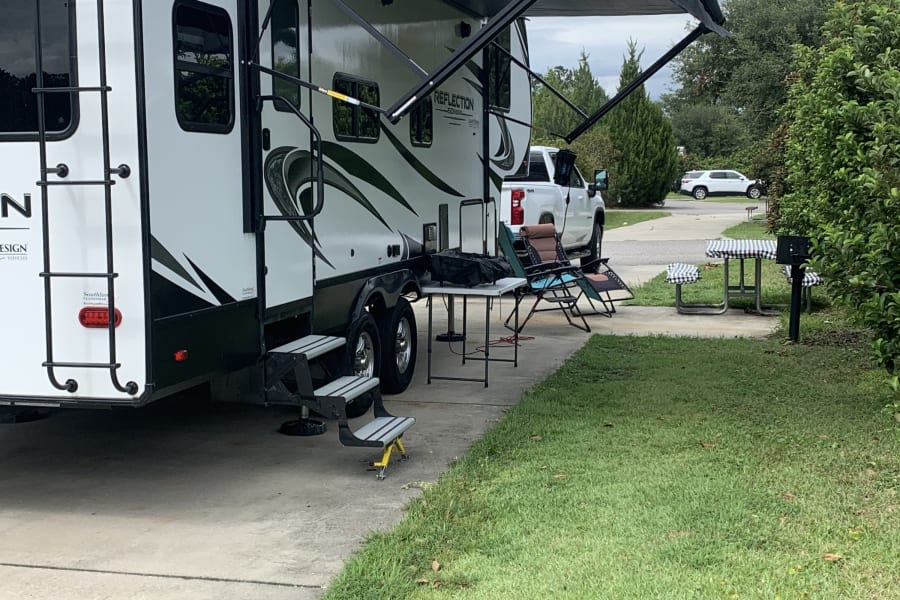 RV rental at a serene campsite: white travel trailer with extended awning, outdoor chairs, and a picnic table on a paved site.