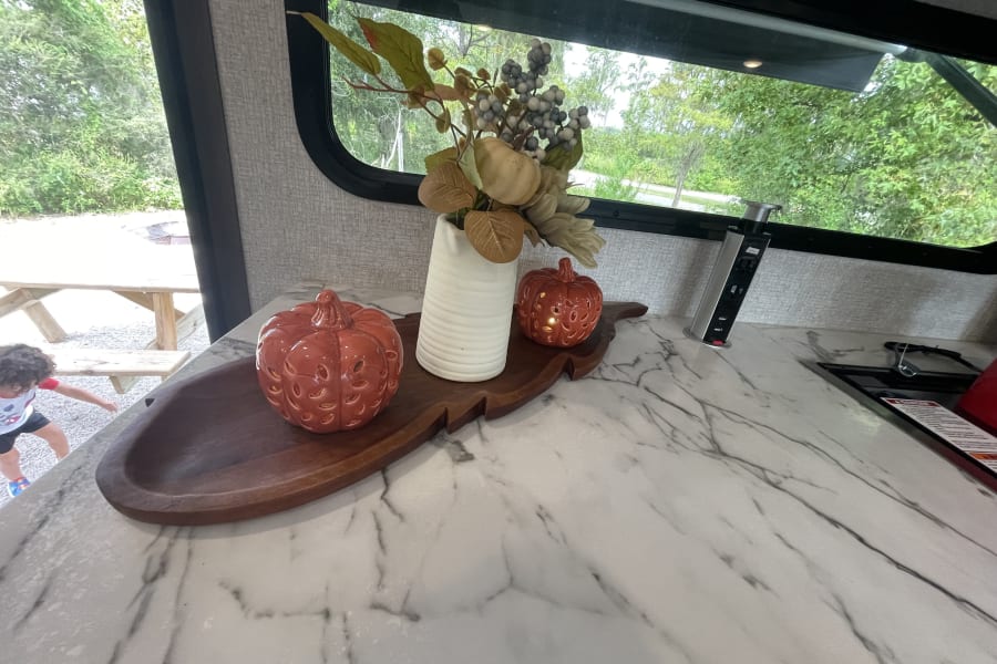 RV rental kitchen: white counter, fall decor, pop-up outlet. Child plays outside near picnic table.