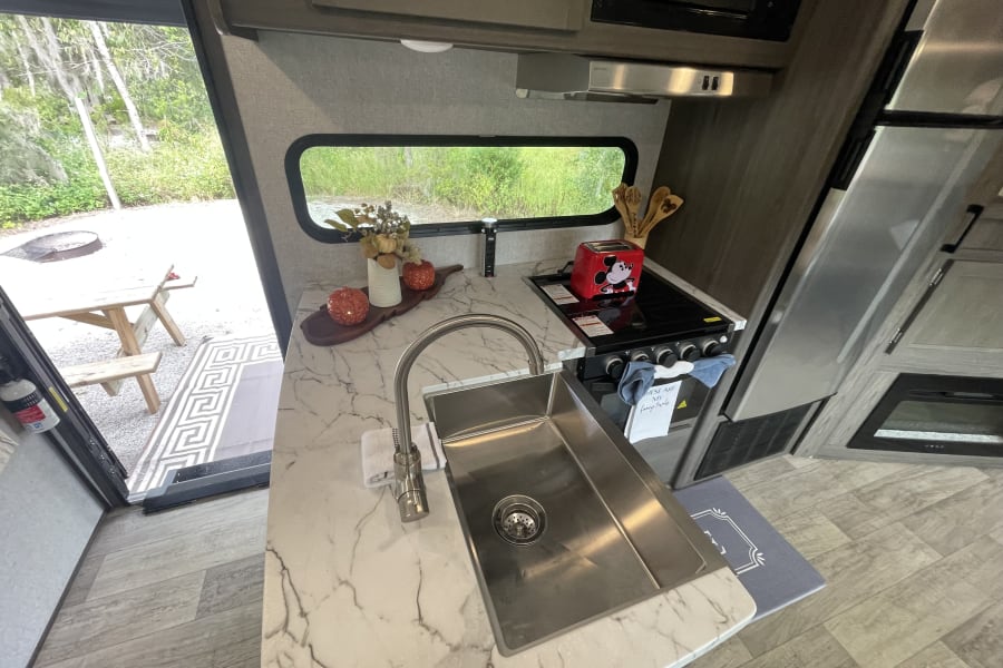 Modern RV rental kitchen with sink, stove, microwave, and festive decor, offering a view of the campsite and picnic area.