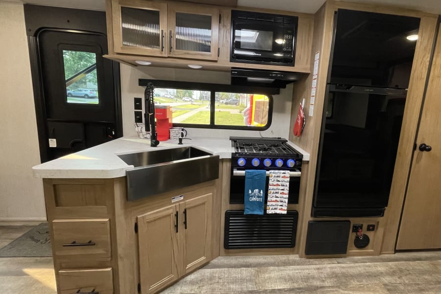 This RV rental features a well-equipped compact kitchen with a full sink, stove, microwave, and fridge, perfect for camping.