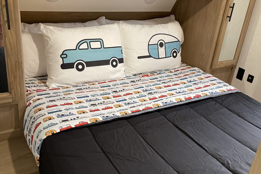 RV rental bedroom features a cozy bed with truck and camper pillows and sheets, plus overhead storage cabinets.