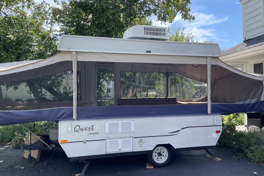 A white and blue Jayco Qwest pop-up RV, fully set up with screen windows, parked outside. Perfect for an RV rental.